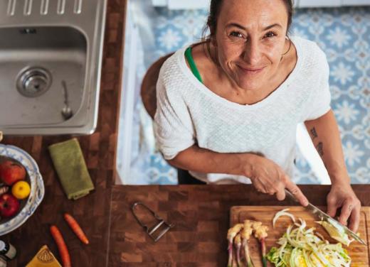 Een vrouw is eten aan het maken en kijkt lachend de camera in